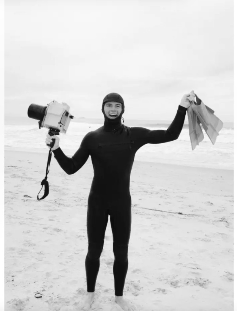 Black and white portrait of Hayden Garfield on a beach in a wetsuit, holding an underwater camera housing and swim fins